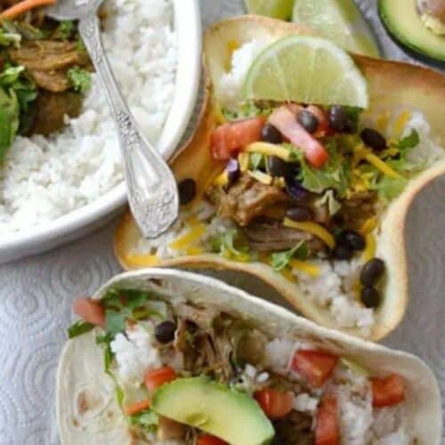 Easy Crockpot Pork Carnitas. Top view of this yummy meal in a baking dish, a soft tortilla and a baked tortilla shell. All the fixings with it, rice, beans, tomato, avocado, cheese, lettuce, tomato, lime