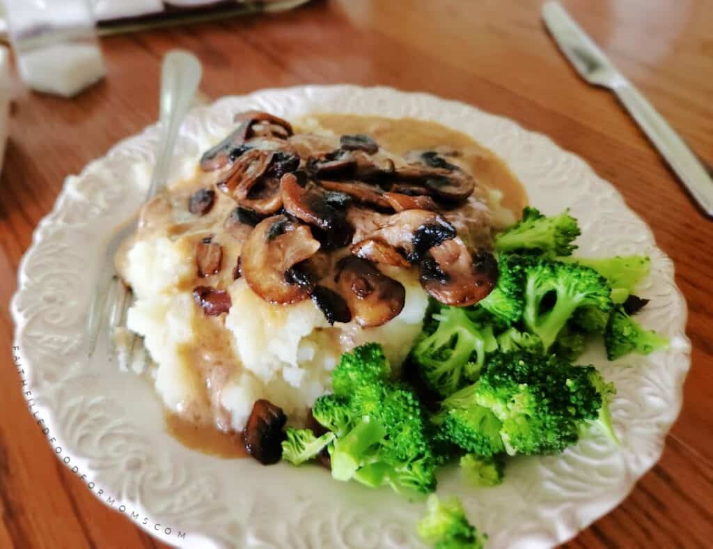 Homemade Salisbury steak with gravy over mashed potatoes, topped with sauteed mushrooms and broccoli on a white plate.