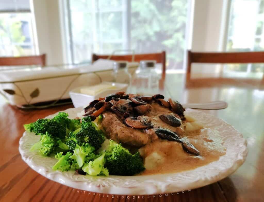 Homemade Salisbury steak on mashed potatoes with gravy and sauteed mushrooms on top and broccoli on the side on a plate.