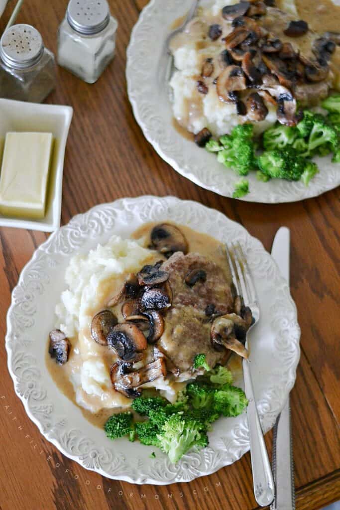Homemade Salisbury steak over mashed potatoes with brown gravy topped with sauteed mushrooms and broccoli on a white plate.