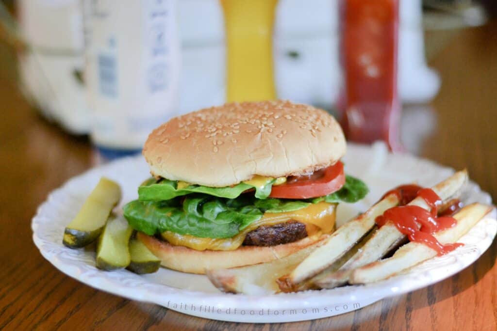 Oven-baked frozen hamburger cheeseburger Juicy oven-baked cheeseburger with lettuce, tomato, and pickles on a sesame bun with fries on a plate. With Steak Fries and pickles on the side.