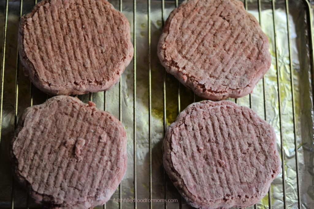 Frozen hamburger patties on wire rack for oven baked hamburgers Frozen hamburger patties arranged on a wire rack over a lined baking sheet before baking.