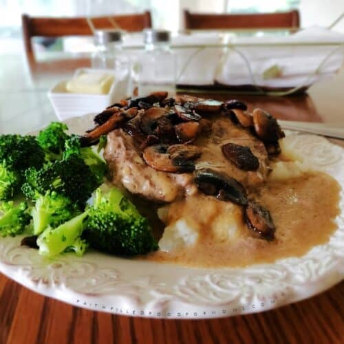 Salisbury steak over mashed potatoes with brown gravy topped with sauteed mushrooms and served with broccoli on a white plate.